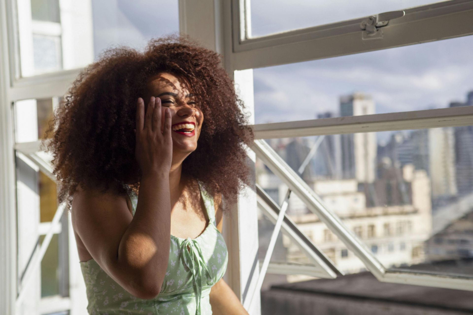 A cheerful woman with afro hair enjoying the sunlight indoors, set against an urban backdrop.